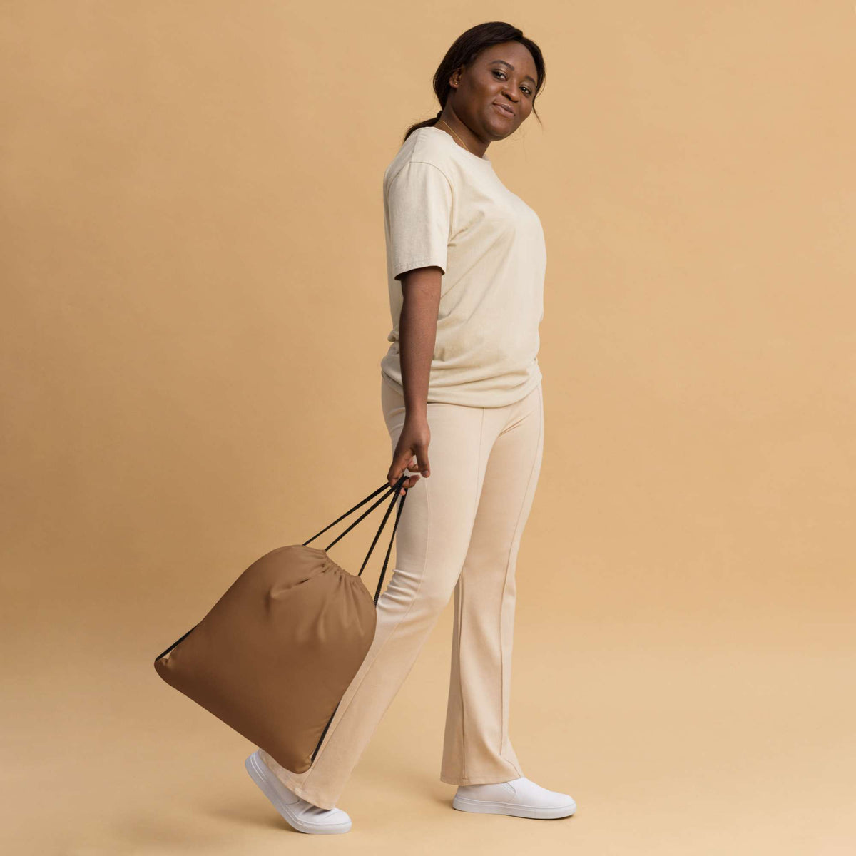 A woman in a cream-colored lounge set walks while carrying a large tan drawstring tote bag against a solid beige studio background. Modern Bold Minimalist Solid Natural Wood Drawstring Bag