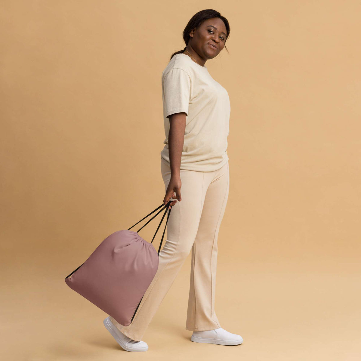 A woman in a cream-colored lounge set walks while carrying a dusty pink drawstring bag with black handles against a solid beige background. Stylish Minimalist Solid Rosy Brown Durable Drawstring Bag