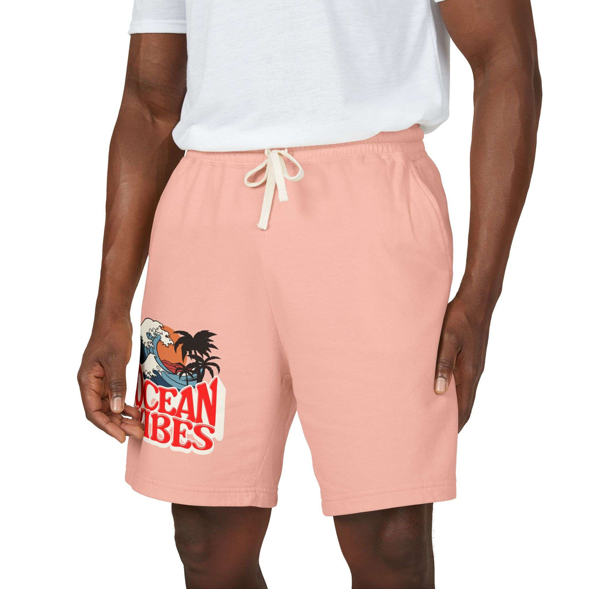 A person wearing peach-pink drawstring shorts with a graphic print on the left leg that reads 'Ocean Vibes' with a tropical wave and palm design, paired with a white t-shirt against a plain white background.
