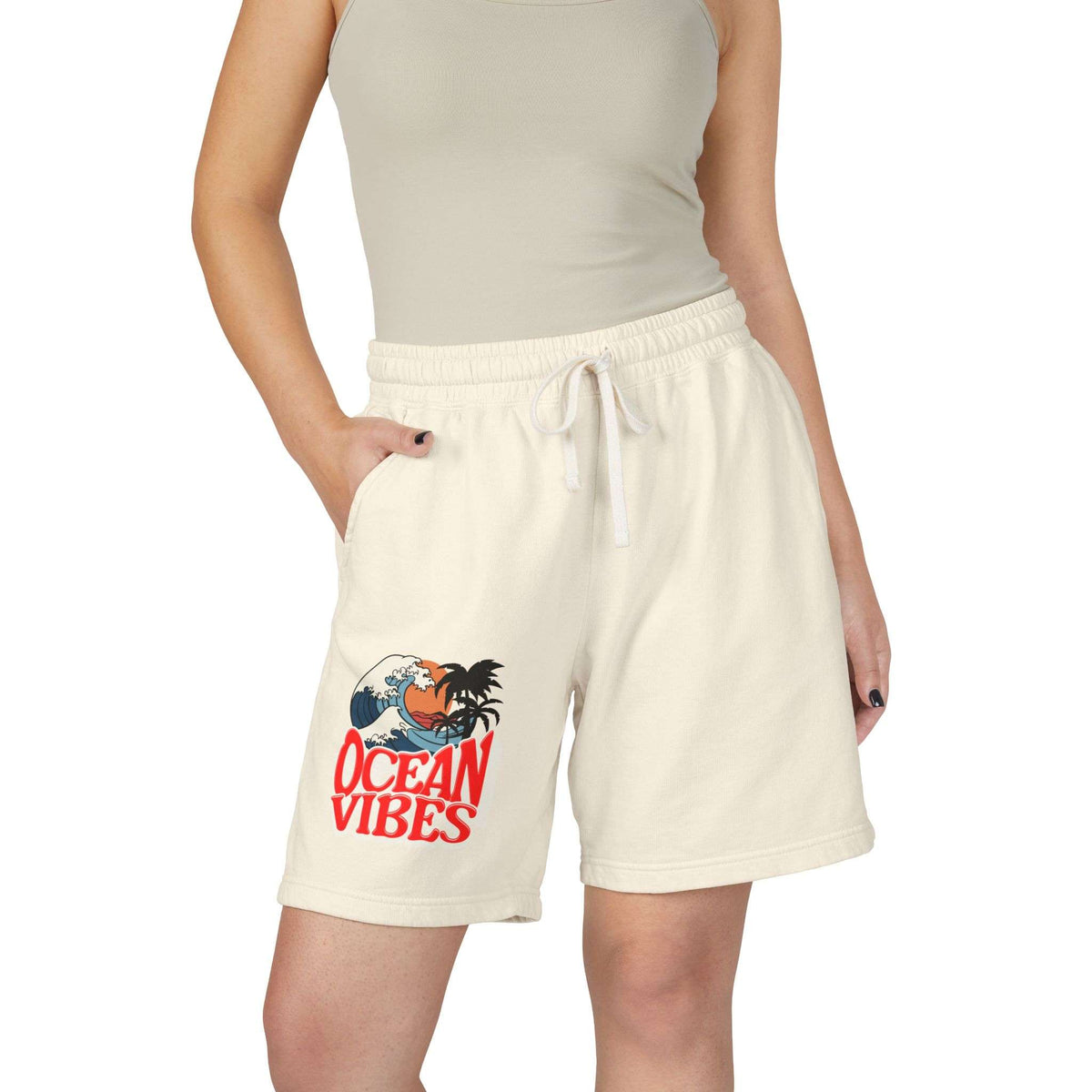 A model wearing cream-colored drawstring shorts with a red and blue 'Ocean Vibes' graphic on the left leg, paired with a beige ribbed tank top against a plain white background. Unique Lightweight Fleece Sweat Shorts in Ocean Vibes Graphic Shop Lightweight Fleece Sweat Shorts in Ocean Vibes for breezy comfort, relaxed style, and an easygoing summer-ready look all day. Refresh your casual rotation with Lightweight Fleece Sweat Shorts in Ocean Vibes—soft, versatile, and ready for sunny days by the shore.