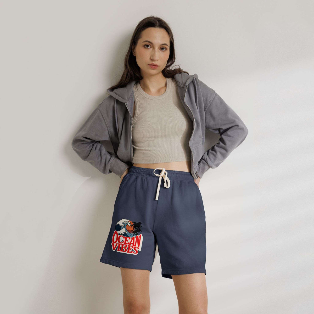 A woman modeling navy blue drawstring shorts with a red and white 'Ocean Vibes' graphic on the left leg, styled with a beige crop top and a gray zip-up hoodie against a light gray wall.