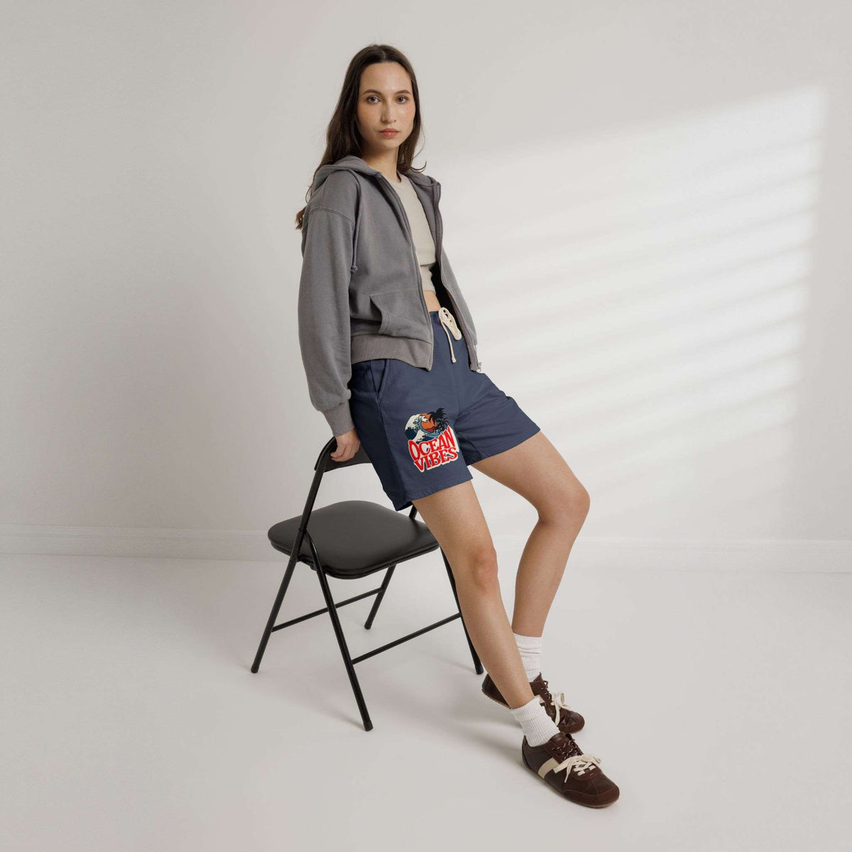 A woman models navy blue drawstring shorts with a colorful graphic print on the left leg, styled with a gray cropped hoodie, white t-shirt, white socks, and brown loafers, while leaning on a black folding chair against a white wall with soft window shadows.