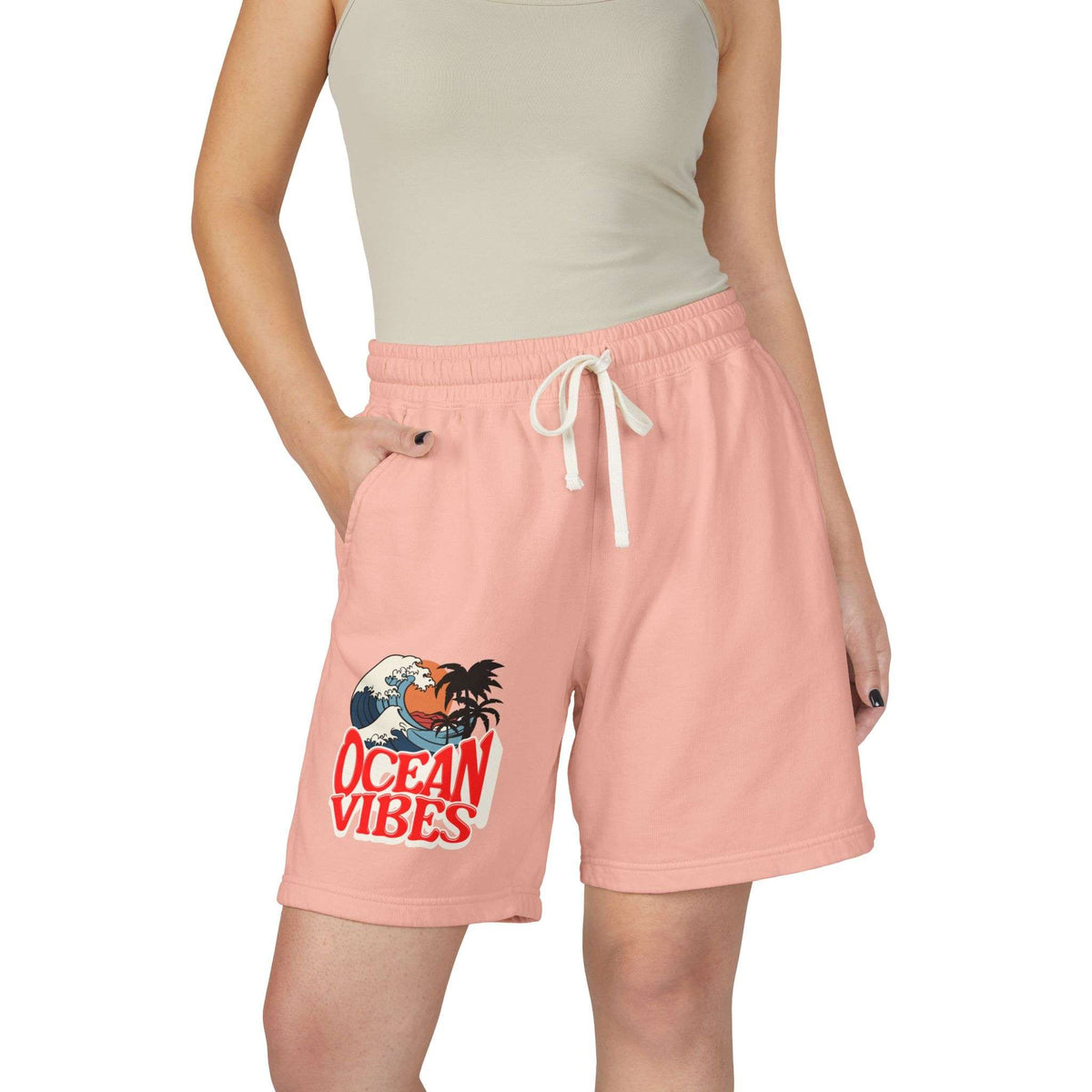A model wearing light peach-pink drawstring shorts with a 'Ocean Vibes' graphic on the left leg, paired with a beige ribbed tank top against a plain white background.