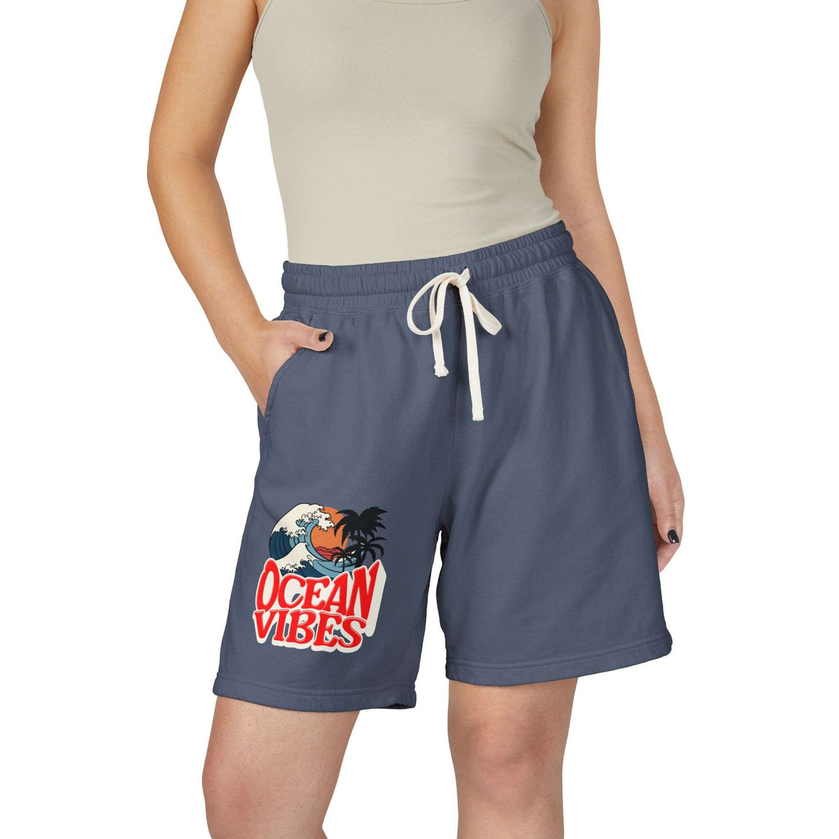 A model wearing slate blue drawstring shorts with a 'Ocean Vibes' graphic on the left leg, paired with a beige ribbed tank top against a plain white background.