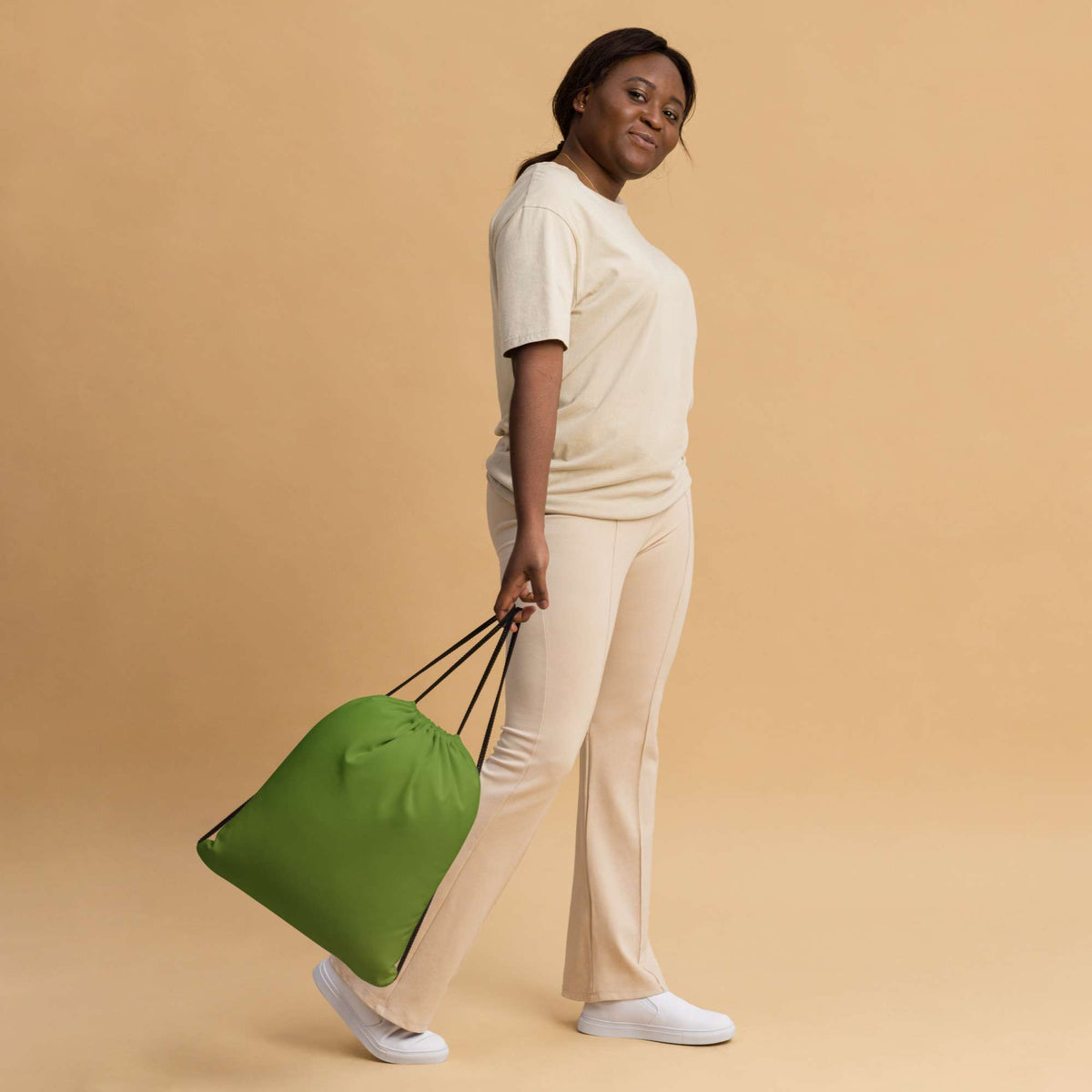 A woman in a cream-colored lounge set walks while carrying a bright green drawstring bag with black handles against a solid beige background. Compact Minimalist Bold Solid Olive Drab Drawstring Bag