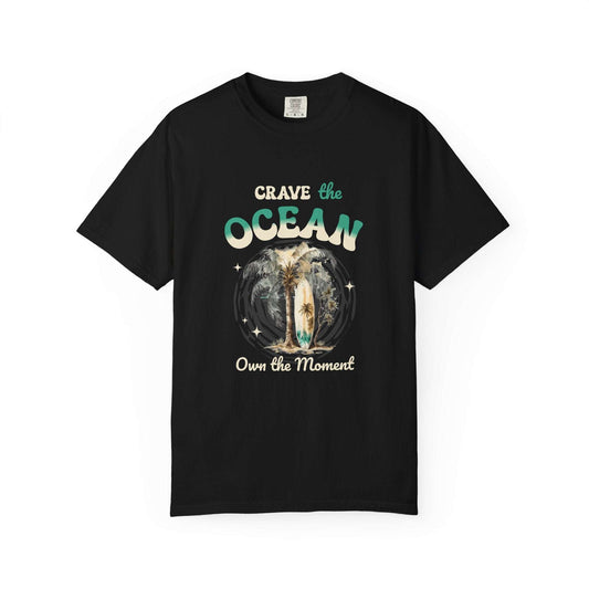A black short-sleeve crewneck t-shirt with a graphic on the front that reads 'Crave the OCEAN Own the Moment' in stylized teal and white lettering, surrounding a central illustration of a surfer on a board. Crave the Ocean Own the Moment Graphic T-Shirt for Casual Wear
