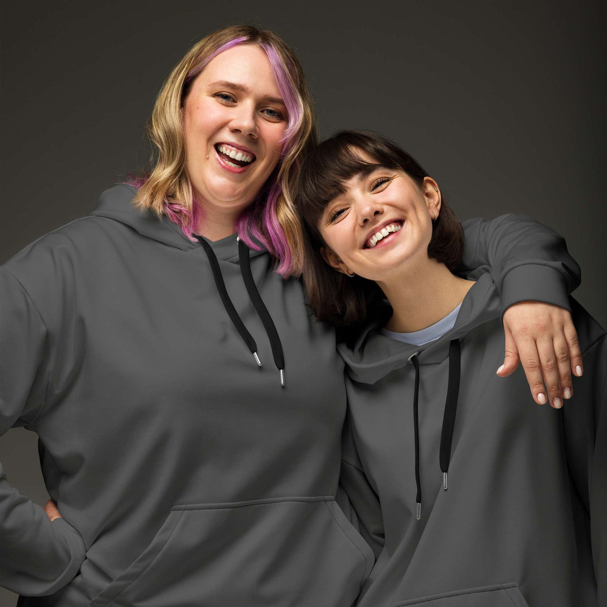 Two women smiling and posing together while wearing matching dark gray hoodies with black drawstrings, photographed against a dark gray studio background. Minimalist Comfortable Plain Gray Hoodie for Relaxed Wear