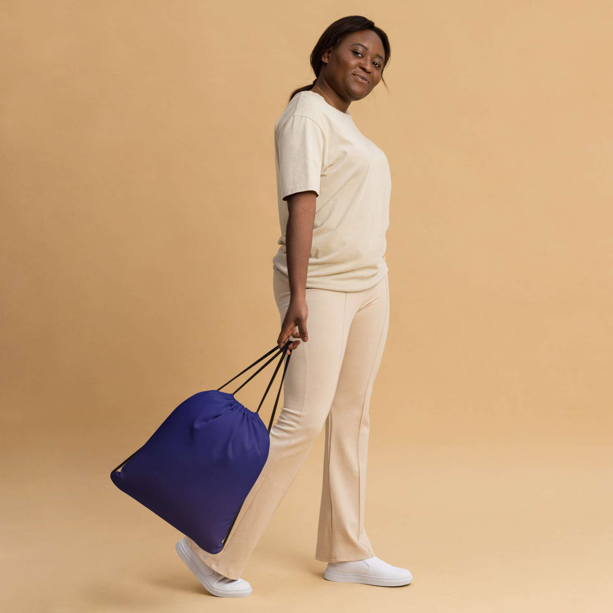 A woman in a cream-colored lounge set holds a navy blue drawstring bag with black handles against a solid beige studio background. Practical Bold Minimalist Solid Midnight Blue Drawstring Bag