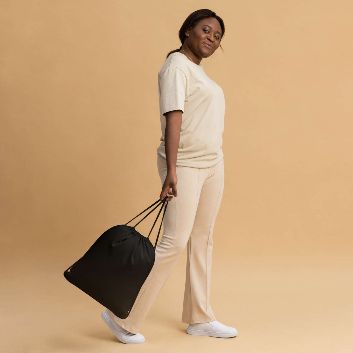 A woman in a cream-colored lounge set walks while carrying a black drawstring bag against a solid beige studio background. Simple Bold Minimalist Solid Black Lightweight Drawstring Bag